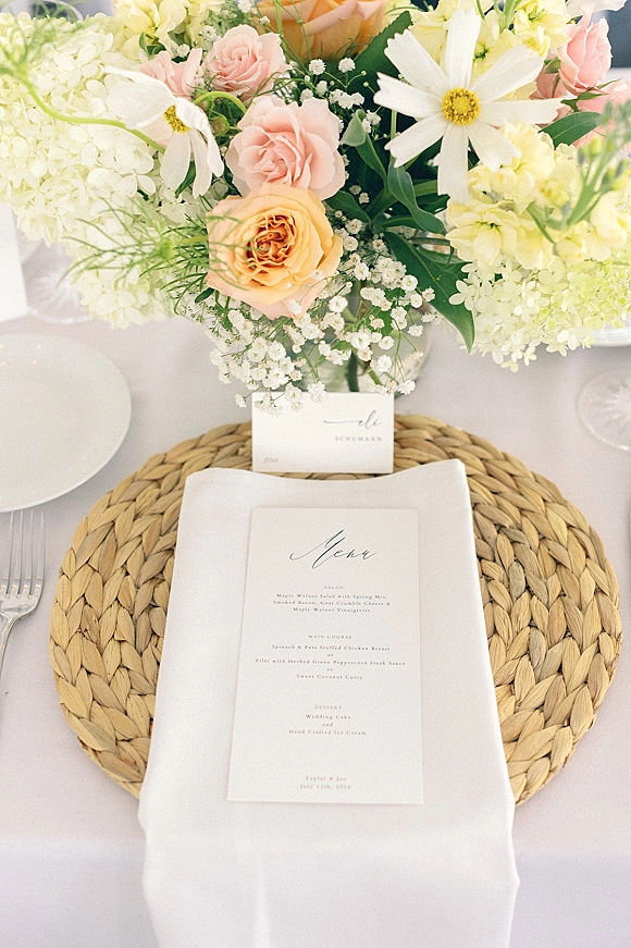 Reception tablescape with wedding table setting, pastel floral centerpiece and menu card atop a woven placemat on white tablecloth