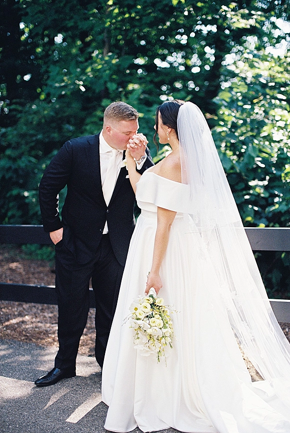 Wedding couple portrait of groom kissing bride’s hand as they hold hands, her long veil and bouquet framed by trees and a wooden fence