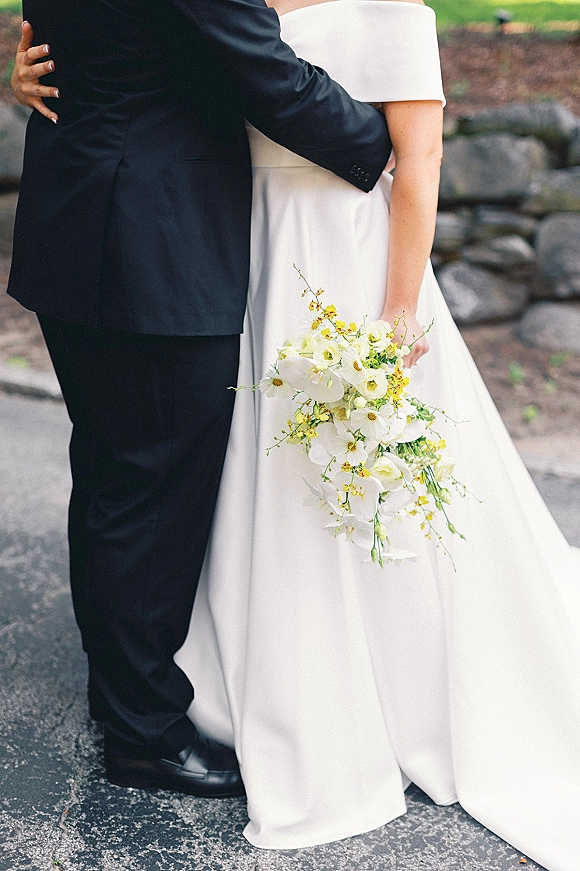 Couple portrait of bride and groom embrace, her white orchid bouquet with yellow accents against a stone wall and greenery path
