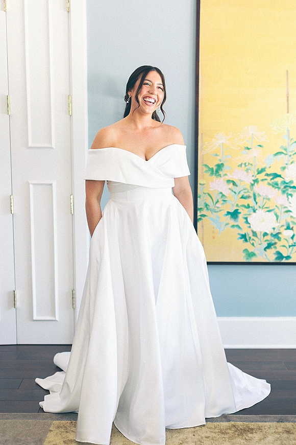 Bridal portrait of a bride in an off the shoulder wedding dress with drop earrings, standing by a blue wall and framed floral art indoors