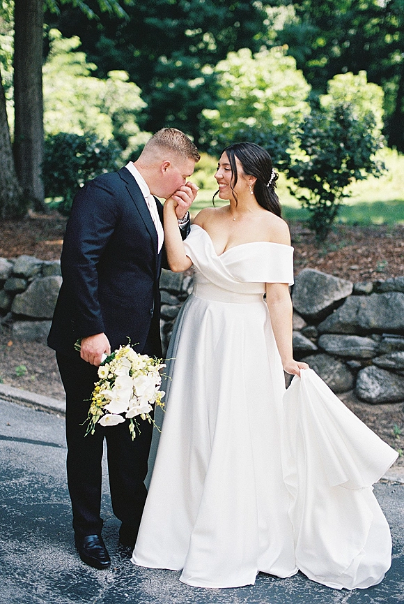 Couple portrait of groom kissing bride’s hand as they hold hands, her off-the-shoulder gown and bouquet by a stone wall path