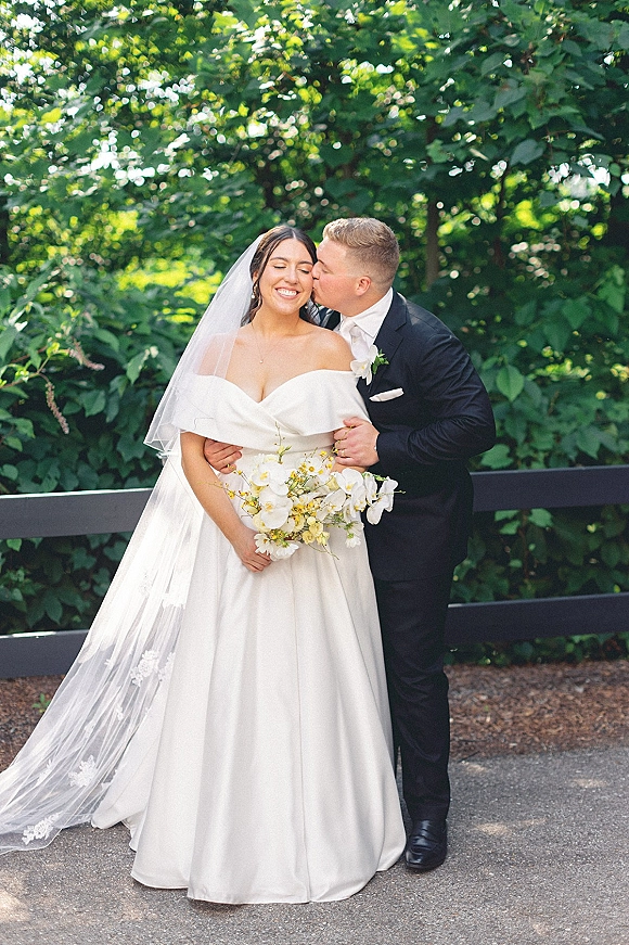 Wedding couple portrait with groom kissing the bride’s cheek as she holds a bouquet, veil trailing against green foliage and a wooden fence