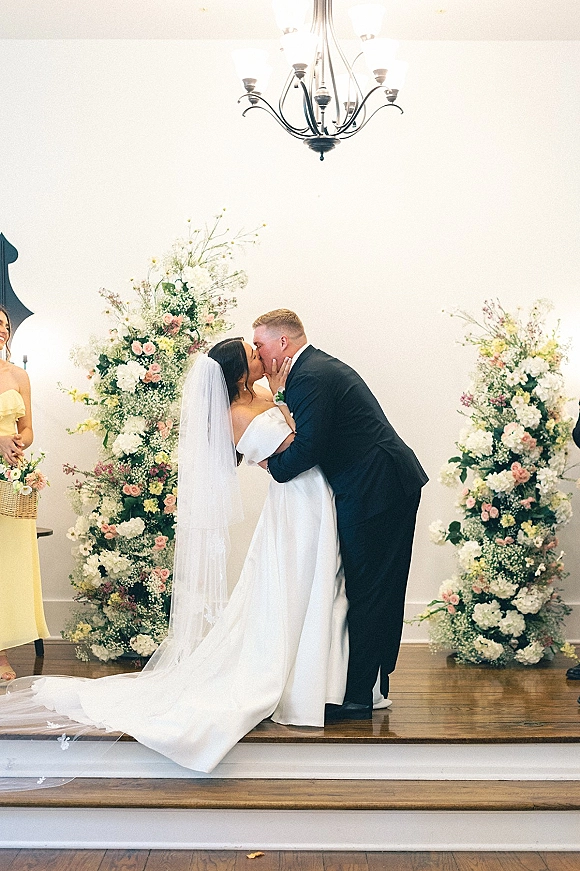 Wedding kiss as the first kiss wedding ceremony, bride in off-the-shoulder gown and veil with groom in suit beneath a chandelier on stage
