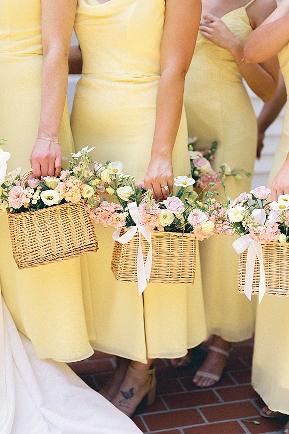 Bridesmaid dresses in butter yellow, shown with wicker baskets of pastel flowers and white bows on a brick floor by a white wall