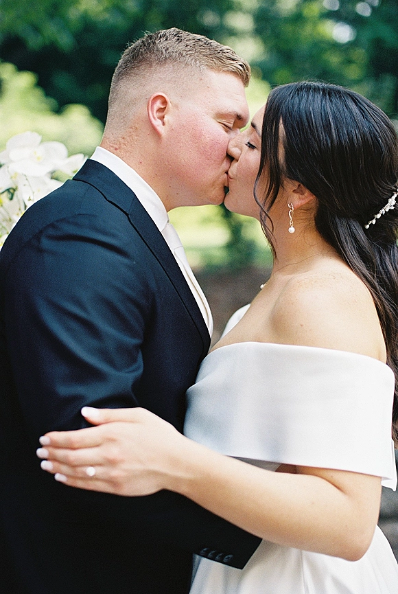 Wedding kiss portrait of bride and groom kissing, bride’s hand with engagement ring on his suit, greenery and trees behind them