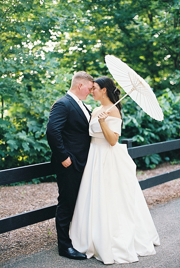 Couple portrait of bride and groom forehead touch, bride holding a white paper parasol on a garden path by green trees and wood fence