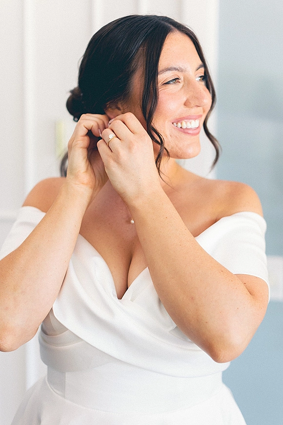 Bride getting ready, bride putting on earrings as window light highlights her off-the-shoulder dress, engagement ring, and pearl pendant necklace