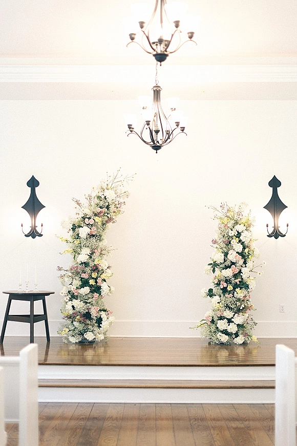 Ceremony altar decor with floral altar pillars of blush and white flowers and greenery on a wooden stage beneath chandeliers, white pews behind