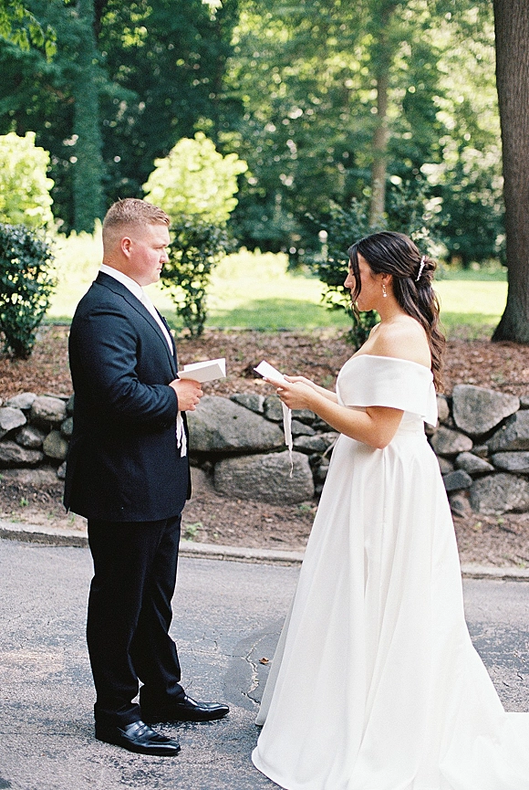 First look moment as bride and groom read paper vows, her off-shoulder wedding dress and hairpiece beside a stone wall path