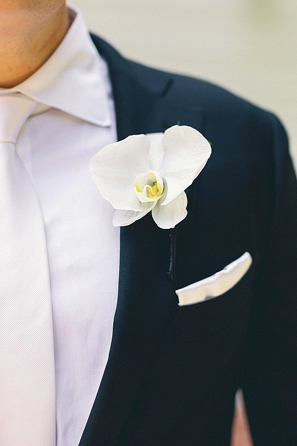 Groom boutonniere with a white orchid boutonniere pinned to a black suit lapel beside a crisp pocket square against a neutral wall