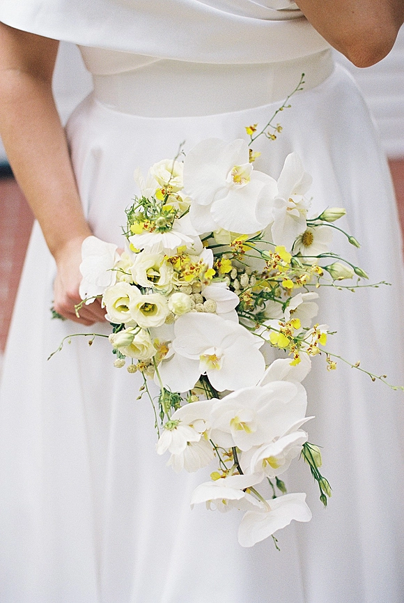 Bridal bouquet with a white orchid bridal bouquet and yellow orchid spray, with daisies, roses, and greenery against a neutral wall in soft indoor light