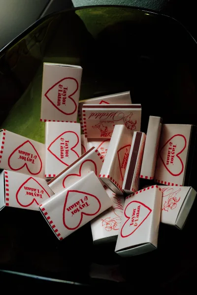 Wedding matchbooks with red heart graphic and bold red typography, personalized names scattered on a black glass surface with green fabric backdrop