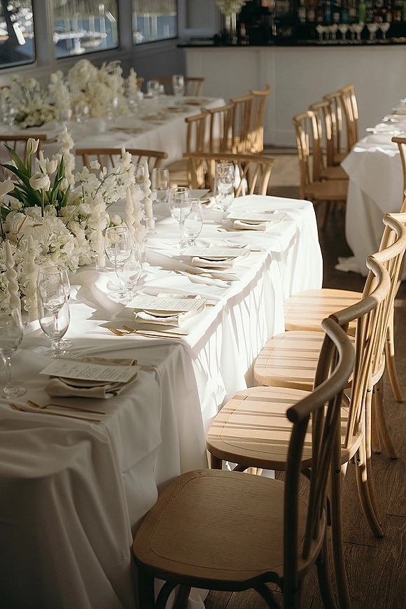 Reception tablescape with a white wedding tablescape of white florals, taper candles, gold flatware, and place cards by bar windows