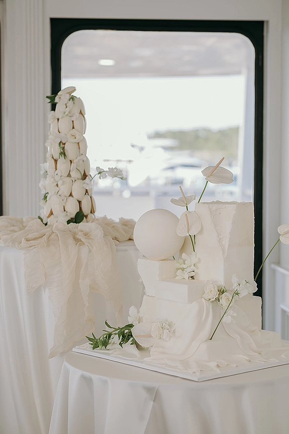Wedding cake with textured buttercream tiers and white anthurium florals beside a macaron tower on a pedestal by a waterfront window