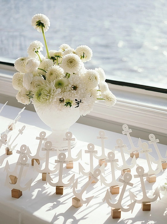 Wedding escort cards in wooden holders with anchor place cards beside a white floral urn on a sunlit windowsill overlooking ocean water