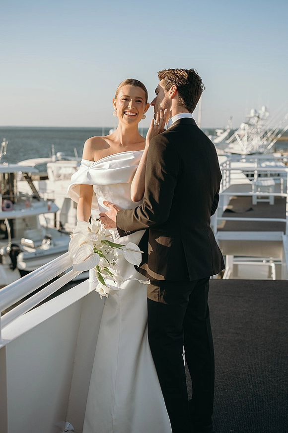 Couple portrait of bride in strapless gown and groom in black tuxedo, cheek kiss by marina yachts, holding anthurium bouquet with orchid accent