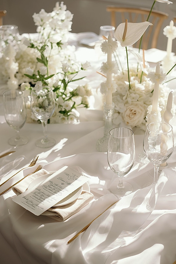 Reception tablescape with white wedding table decor, anthurium and rose centerpiece, taper candles in clear glass, gold flatware, menus in sunlit room