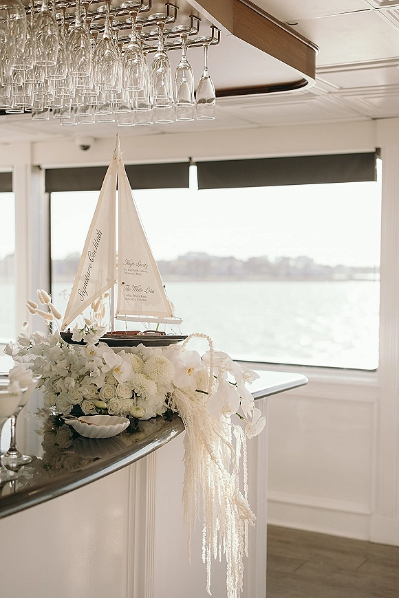 Wedding bar decor with a bar top floral arrangement of white roses and orchids, hanging wine glasses, and a sailboat by waterfront windows