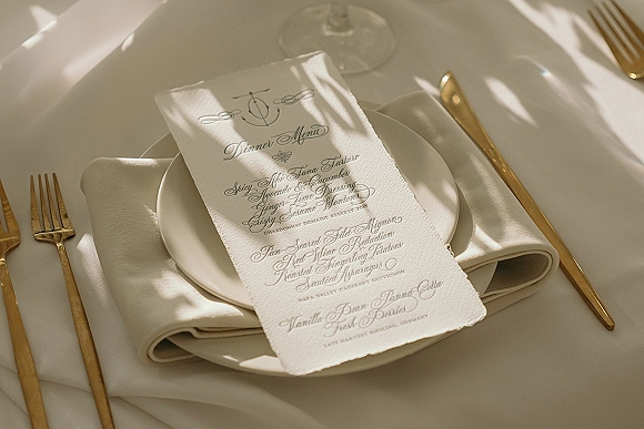 Wedding menu card with calligraphy wedding menu on deckle edge paper atop a white plate, ivory napkin, gold flatware in dappled sunlight