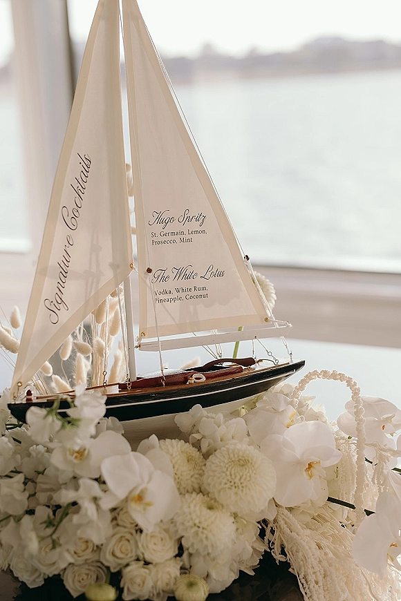 Signature cocktail menu displayed beside a sailboat model with white florals and beaded garland, lit by window light with ocean view