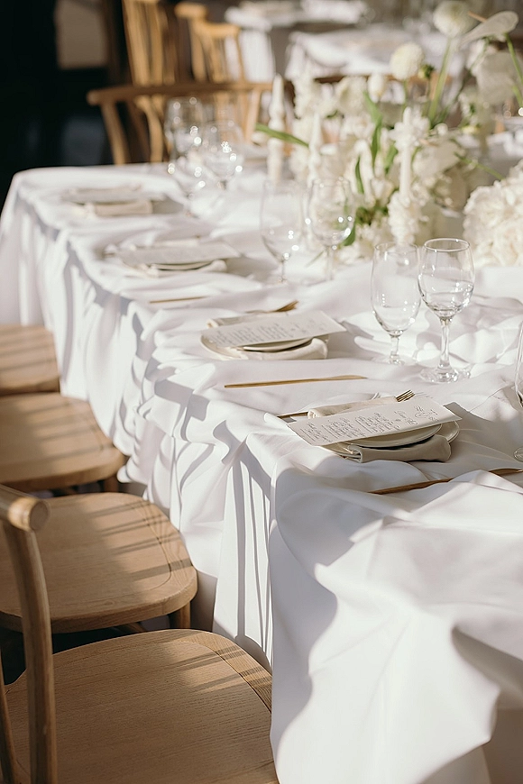 Reception tablescape with a white wedding tablescape of gold flatware, menu cards, wine glasses, white florals, and tapered candles in sunlight shadows