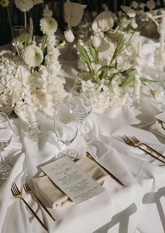 Reception tablescape with a white wedding tablescape of tulips and anthurium, twisted taper candles in glass, and gold flatware on linen