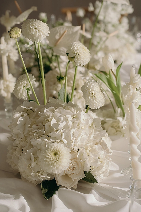 Wedding floral centerpiece of white hydrangeas, roses, and tulips with taper candles in glass holders on white linen, reception tables blurred behind