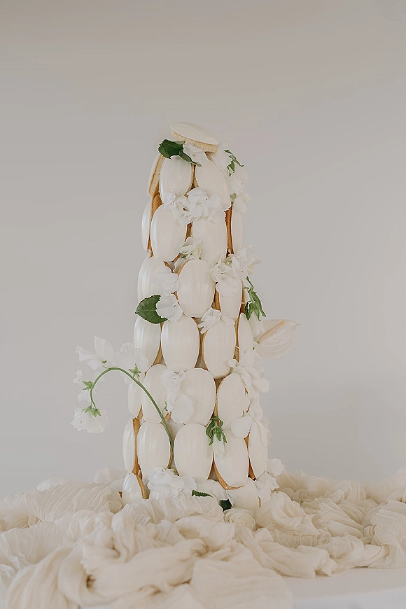 Wedding cake with macaron wedding cake tower, topped with white flowers and greenery, set against neutral draped fabric backdrop