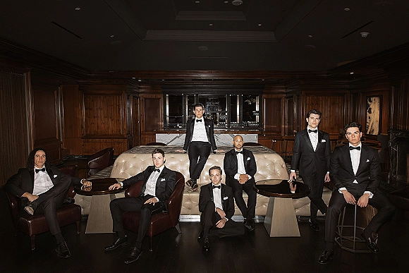 Groomsmen portrait in black tuxedos with bow ties, seated and standing around leather lounge chairs in a wood-paneled bar setting