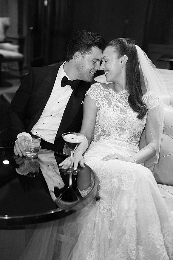 Couple portrait in a dark lounge, bride in lace dress and veil leaning into tuxedoed groom on sofa, holding cocktails on glass table