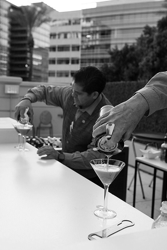 Wedding cocktail bar with signature cocktail station featuring martini glasses and shaker on an outdoor terrace with palm and buildings behind