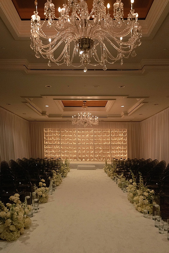 Ceremony setup with a white aisle runner lined with low florals and glass cylinder candles beneath crystal chandeliers in a ballroom