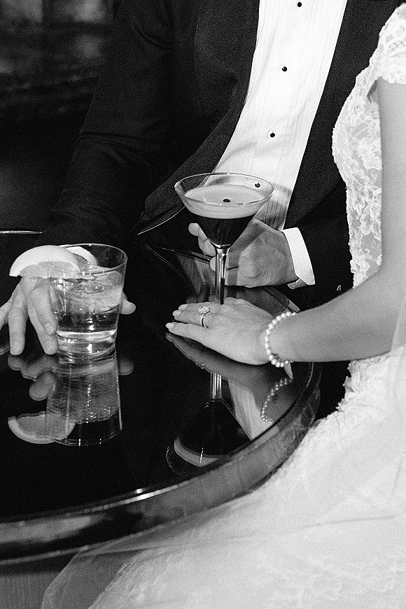Wedding toast with bride and groom hands holding cocktail glasses, engagement ring and lace dress detail on a mirrored table in a dim lounge setting