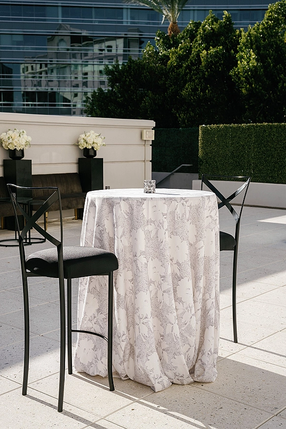 Cocktail table setup with patterned high-top tablecloth, white floral arrangement and votive candle, plus black cross-back chairs on terrace patio
