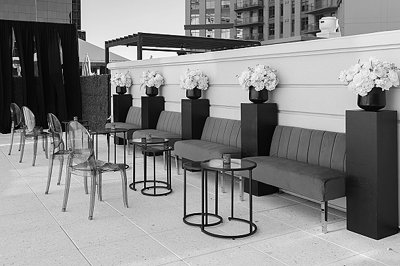 Wedding lounge setup with an upholstered sofa, clear acrylic chairs, and round cocktail tables on a rooftop terrace with black drape backdrop