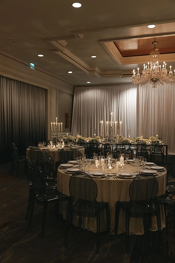 Reception tablescape with candlelit wedding reception glow, beige linens, white floral centerpieces, and crystal chandelier over white drape backdrop
