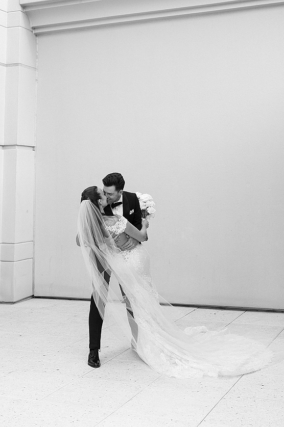 Wedding kiss portrait of bride and groom kissing in a dip, her veil blowing as he holds her by a white wall with column