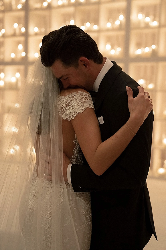 Wedding first dance as bride and groom dancing in a close embrace, her cathedral veil trailing over a lace dress beneath string lights indoors
