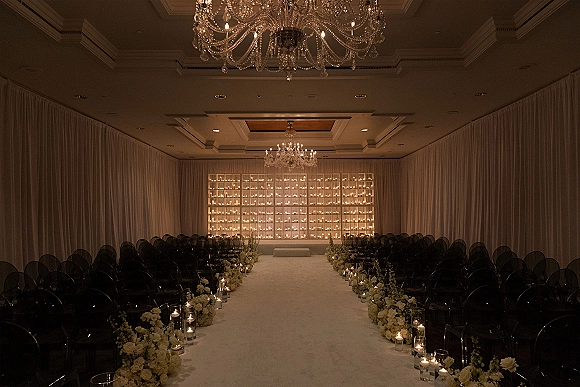 Indoor ceremony setup with a ballroom wedding ceremony white aisle runner, pillar candles in glass holders, black chairs, and draped chandeliers backdrop