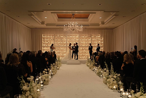 Wedding ceremony with bride and groom on a raised stage, white aisle runner lined with florals and candlelight in a ballroom setting