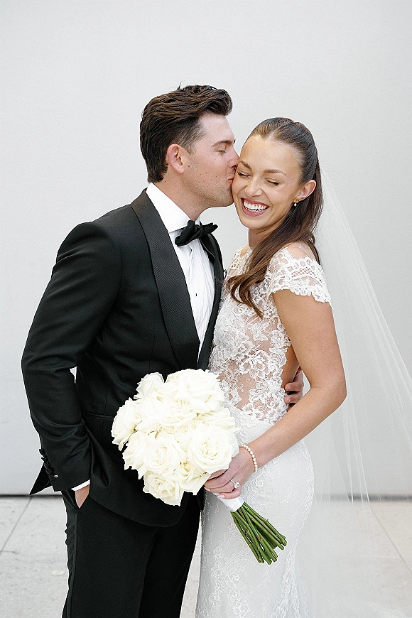 Wedding couple portrait with groom kissing bride’s cheek as she laughs, holding a white rose bouquet against a white wall backdrop