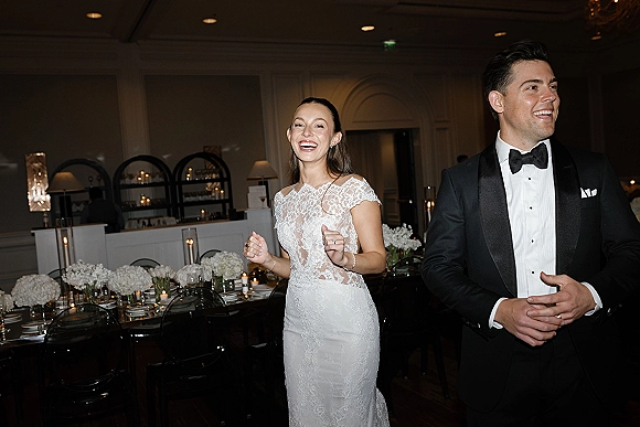 Reception entrance as bride in a lace wedding dress laughs with groom in tuxedo beside a long candlelit banquet table indoors