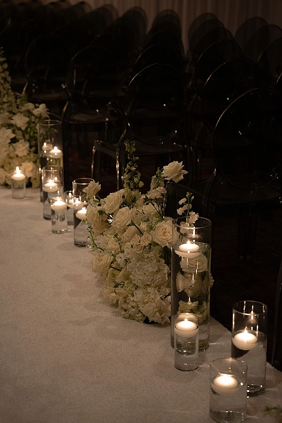 Ceremony aisle decor with wedding aisle candles in glass cylinders, white roses and hydrangeas lining a white runner in a dim hall