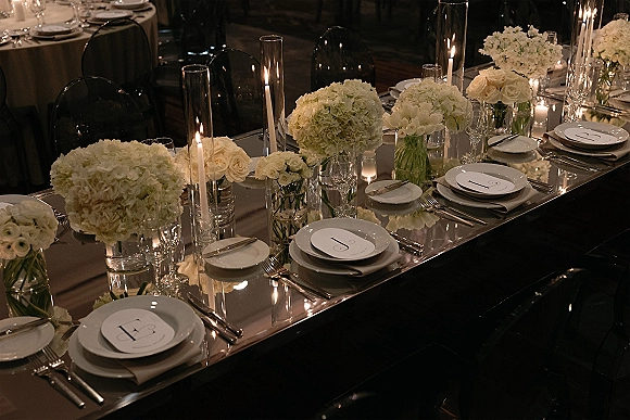 Reception tablescape on a mirror wedding table with white hydrangea and rose centerpieces, tall taper candles, and monogram plates in dim lighting