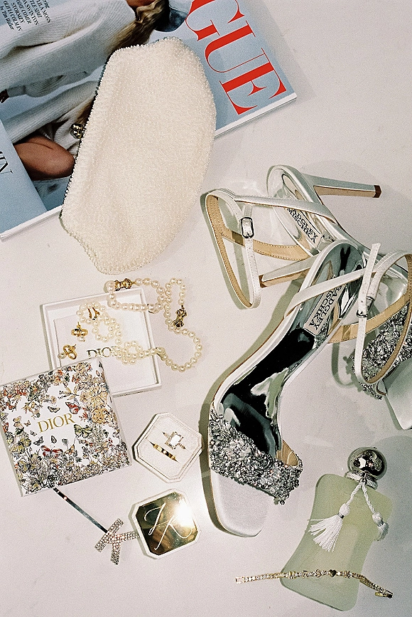 Bridal accessories flatlay with rhinestone high heels, pearl clutch and necklace, rings in boxes, and perfume bottle on white background