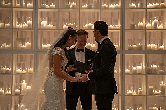 Wedding vows as bride in lace dress and veil holds groom’s hand while officiant reads beside candles on a candle wall backdrop