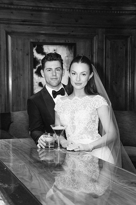 Couple portrait in a bar booth, bride in lace wedding dress and veil with groom in tuxedo, both holding cocktail drinks by wood paneling