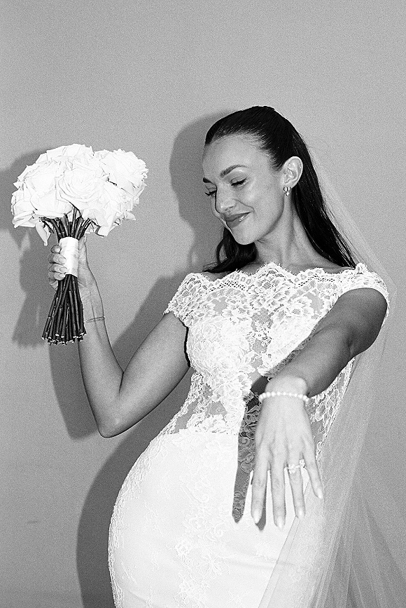 Bridal portrait in black and white of a bride in a lace wedding dress with long veil, holding a rose bouquet and showing her ring by a plain wall