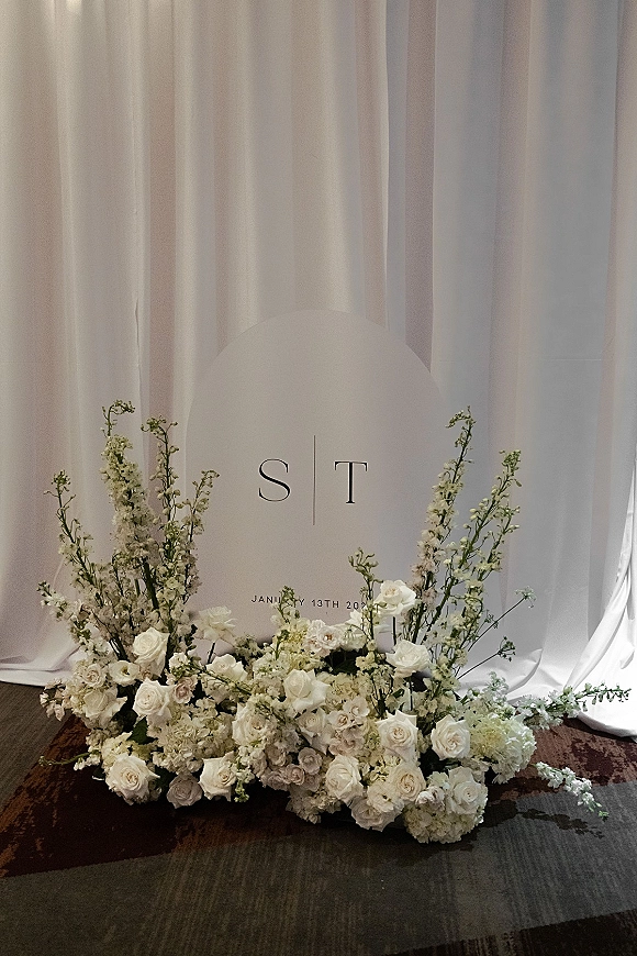 Wedding welcome sign with monogram lettering on a round wedding sign, framed by white draping and a white rose floral base on carpet