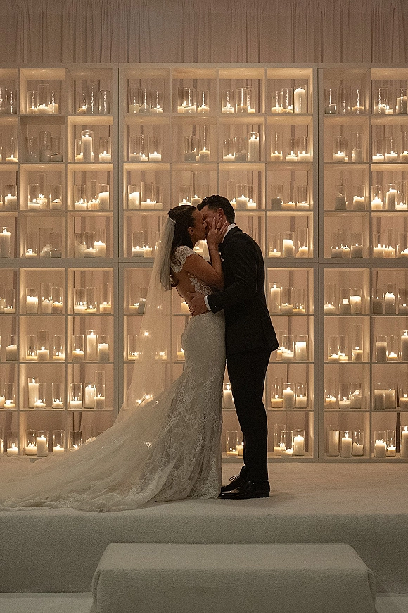 Wedding kiss portrait of bride and groom kissing, her lace dress and long veil flowing beside candlelit glass hurricane vases on white shelves